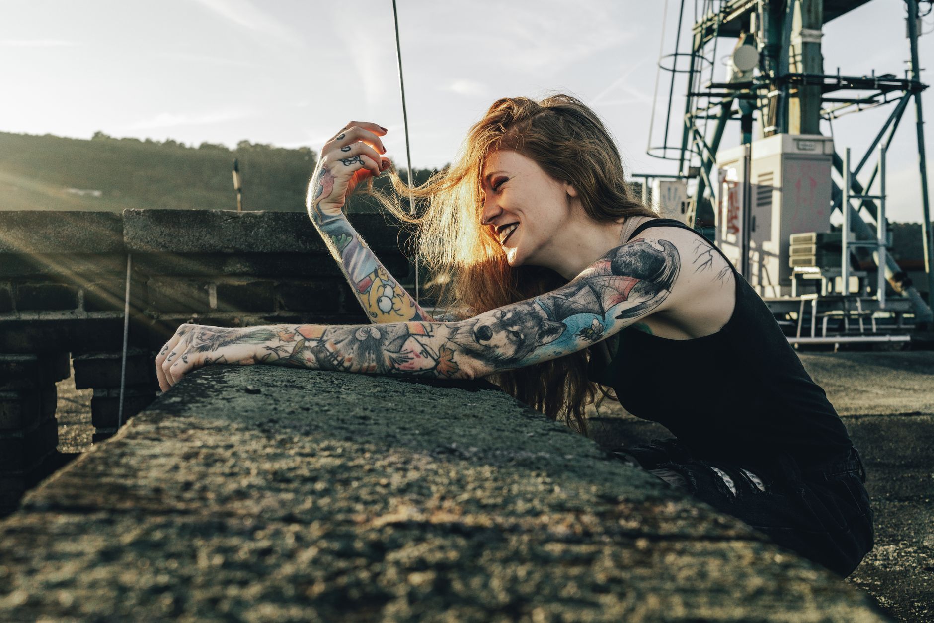 Femme aux bras tatoués souriant, assise sur un toit-terrasse urbain